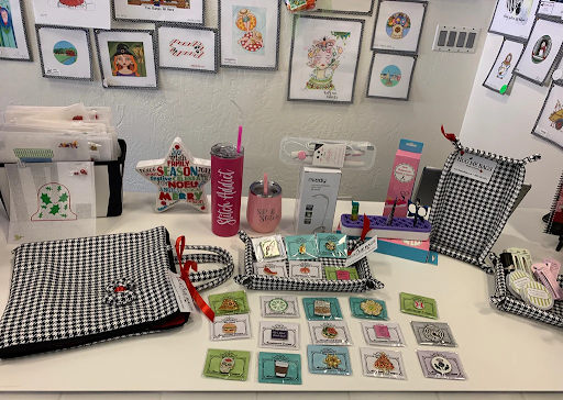 Table with needlepoint designs, accessories, and stitched items, showing the variety offered by modern needlepoint shops online.