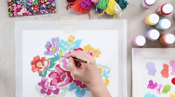  Hand painting flowers on a needlepoint canvas with threads and a reference design nearby using beginner accessories.